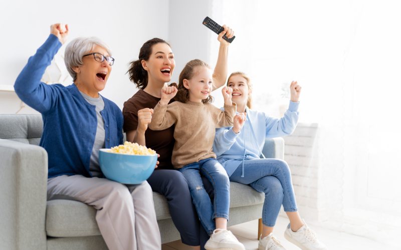 Happy family watching TV, movies with popcorn at home. Mother, daughters and grandmother spending time together.